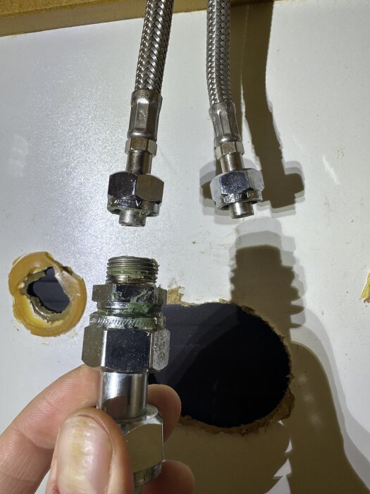 Vattenanslutning med rak koppling och grönt vax på soft-pex-rör, hålls av en hand, omgiven av andra rör och hål i bakgrunden.