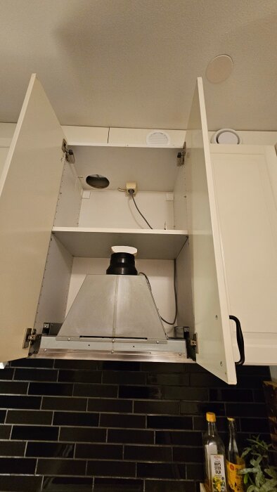 Köksfläkt installerad i ett skåp, med öppna dörrar, exponerar fläktens anslutning till ventilation. Två flaskor syns på diskbänken bredvid fläkten.