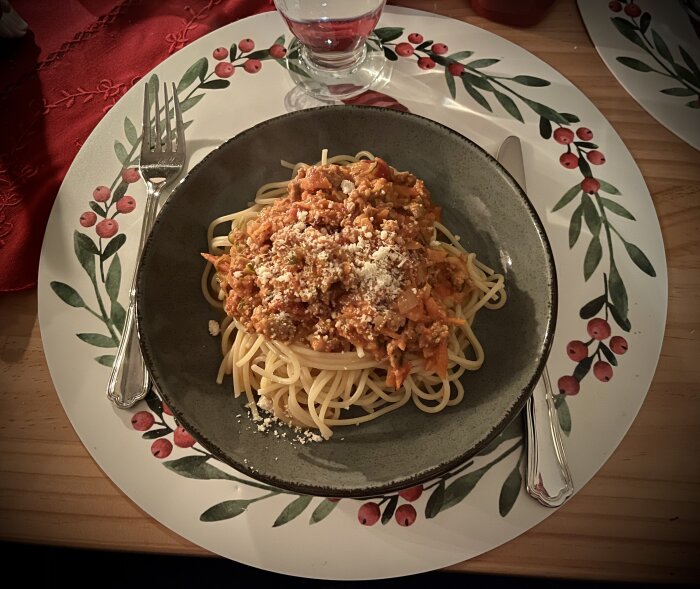 Spaghetti med köttfärssås toppad med parmesan, serverad på dekorerad tallrik, med bestick och ett glas vatten intill.
