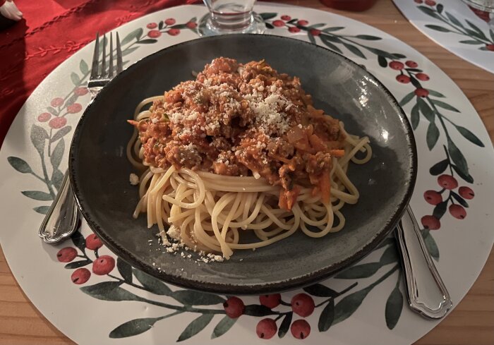 Tallrik med spaghetti och köttfärssås, dekorerad med riven ost, placerad på vit tallrik med rött och grönt bladmotiv.