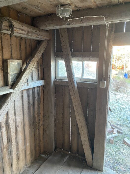 Utomhusvy av en delvis öppen, äldre träkonstruktion med synliga bjälkar och ett fönster.