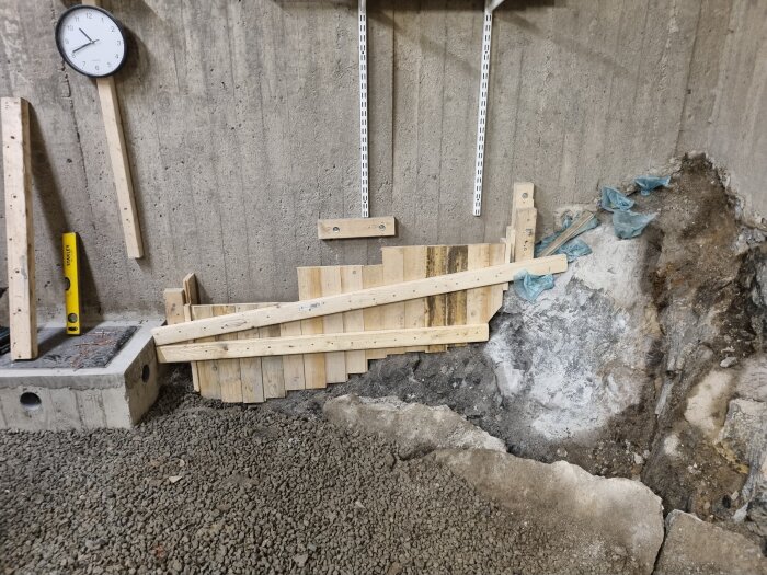 Byggarbetsplats med form för betonggjutning, träslana som stöd, klocka på väggen och stenkross på marken.