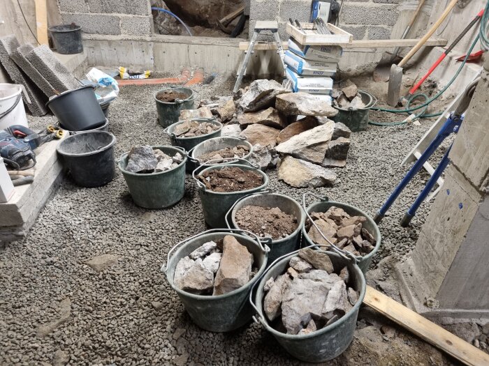Hinkar fyllda med sten och jord står på ett grusgolv inomhus, omgivna av byggmaterial och murade väggar i ett byggprojekt.