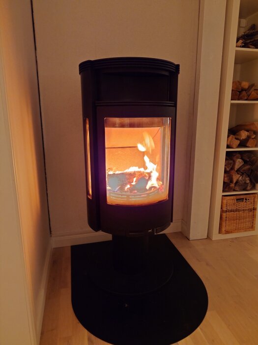 Svart modern kamin med eld inuti, placerad i hörnet av ett rum med trägolv. Vedförvaring syns i hyllan intill.