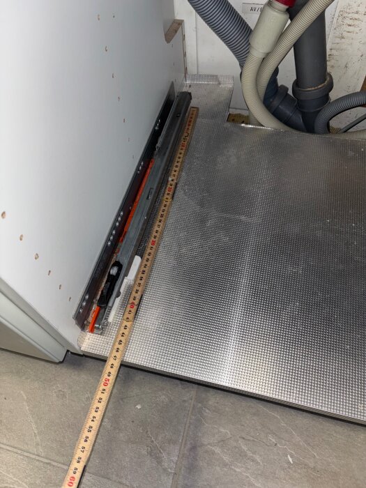 Köket visar en lådkena under diskhon, som är böjd och orsakar att lådan slår i diskmaskinen. Ett måttband ligger på golvet.