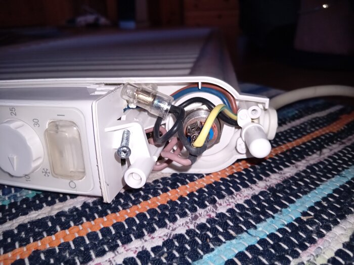 Elektrisk radiator med öppen termostat och exponerade kablar, liggande på en randig matta.