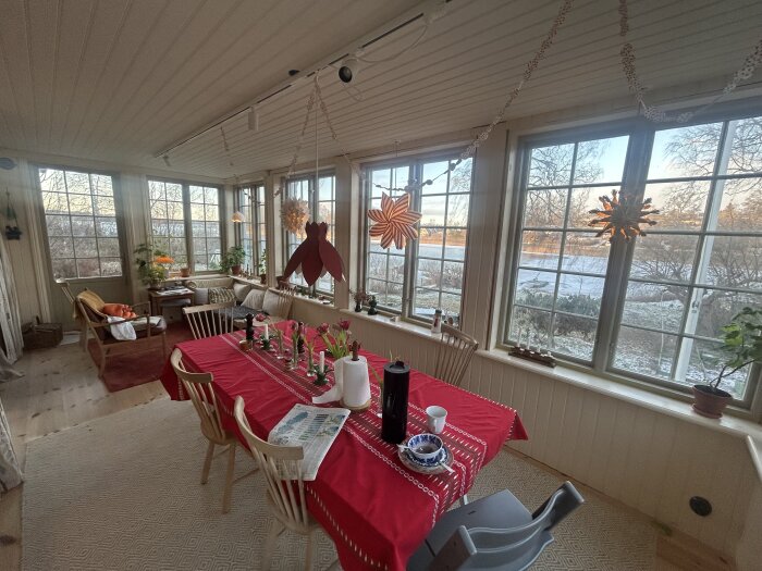 Inglasad veranda med rött dukat bord och blomdekorationer, stolar av trä, stora fönster med utsikt över snötäckt landskap och dekorationer i fönstren.