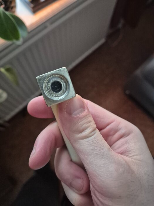 Hand håller ett metallfönsterhandtag med en fyrkantig ände, upplevs lös och snurrar vid försök att öppna, i ett rum med gröna växter.