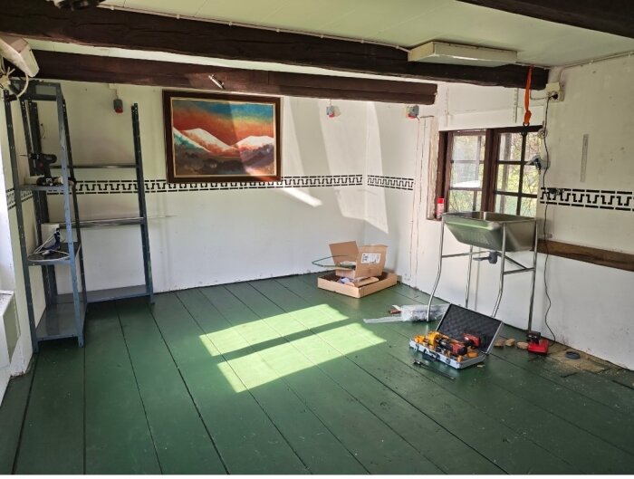 Renoverat rum i torp från 1850 med grönmålade breda plankgolv, verktyg på golvet, en hylla och diskbänk, samt ett fönster och en tavla på väggen.