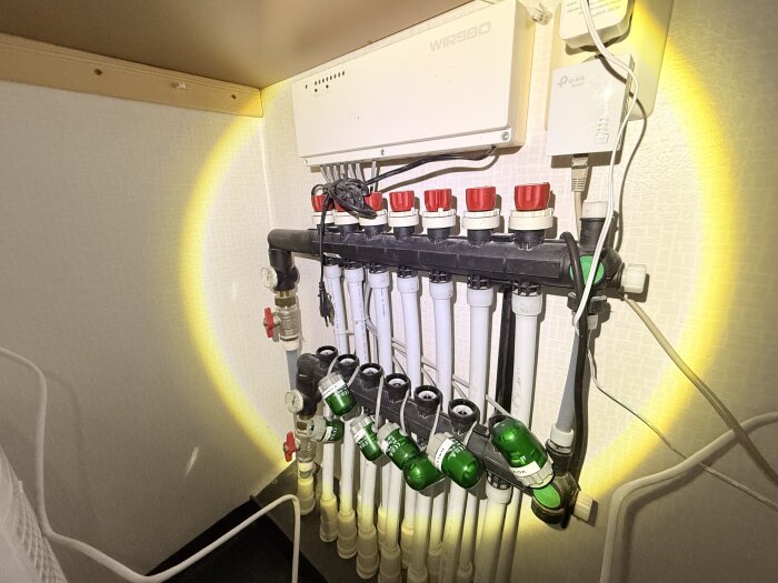 Golvvärmefördelare med flera anslutna slingor, där ett stift sitter lägre än övriga. Fördelardonet är i plast och upplyst av ficklampa.