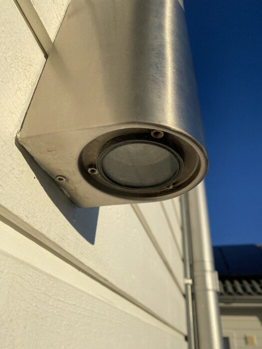 Utomhusvägglampa i rostfritt stål monterad på vit träfasad, med blå himmel i bakgrunden.