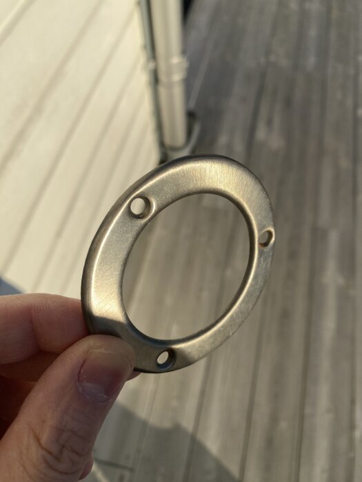 En hand håller en metallring med tre hål, troligen en del av en byggbeslag, mot en bakgrund av trä och en stupränna.