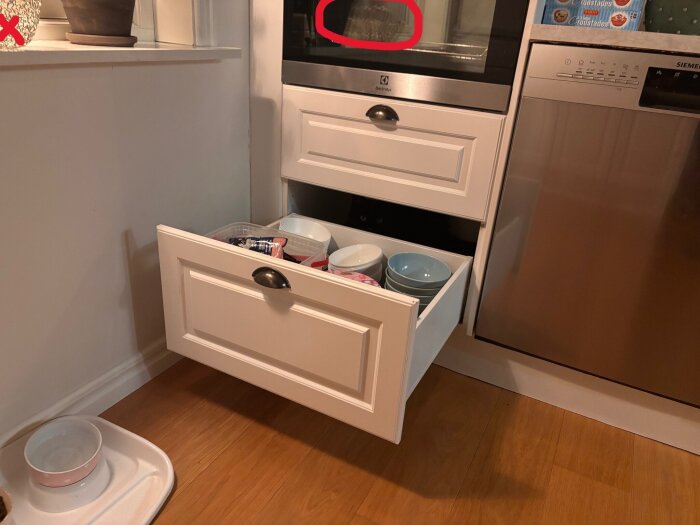 Öppen kökslåda med skålar och förpackningar, vit ugn och diskmaskin i rostfritt stål bredvid. Skål för husdjur på golvet.