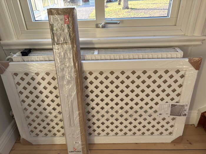 Förpackat vitt elementskydd med rutmönster står lutat mot fönster och radiator på trägolv.