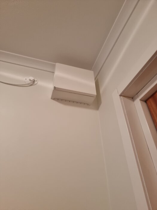 Väggmonterad vit ventilationskanal nära taket i ett hörn av ett rum med vita väggar och en del av en dörrkarm synlig.