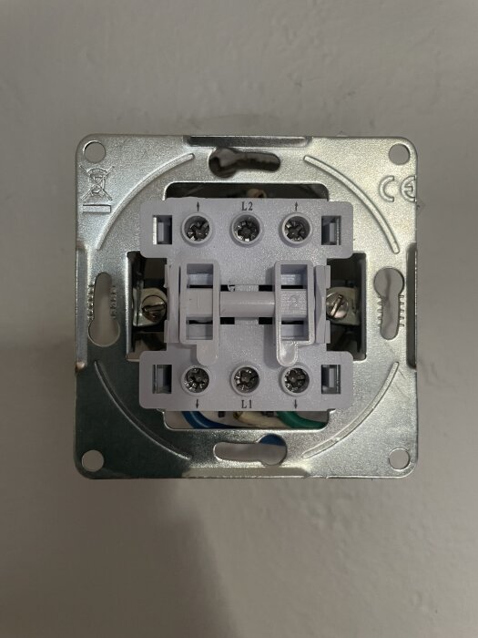Närbild av baksidan på en lampknapp med två brytare, märkta L1 och L2, monterad i en metallram för vägginstallation.