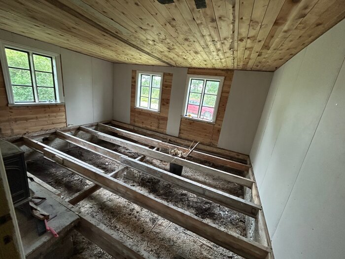 Öppet fritidshusgolv med synligt bjälklag, omgiven av träpanel och fönster, redo för isolering med stenull enligt renoveringsplanen.