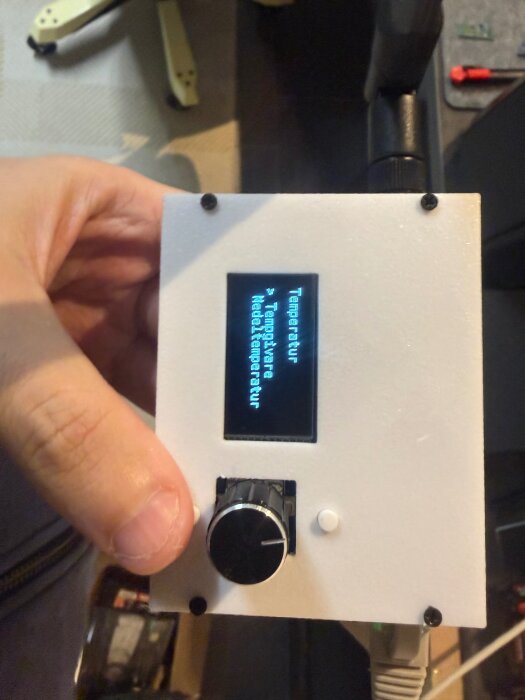 En hand håller en vit elektronikenhet med en liten skärm och en ratt. Skärmen visar text relaterad till temperatur och batterinivå.
