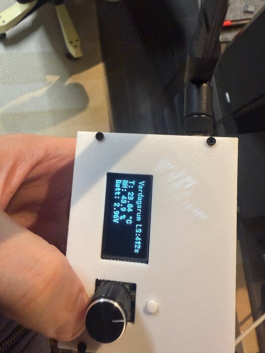 En hand håller i en elektronisk enhet med en skärm som visar temperatur, luftfuktighet och batterinivå. Enheten har en antenn och en ratt.