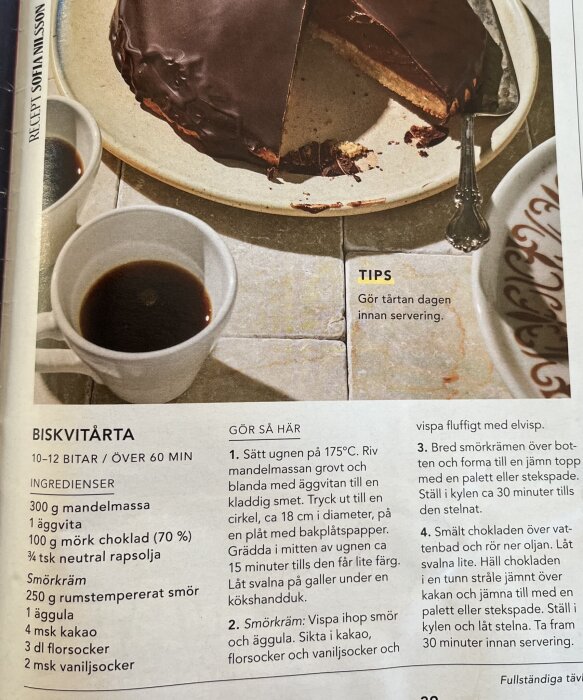 Biskvitårta med ett snitt, serverad på en tallrik bredvid två koppar kaffe och receptbeskrivning.