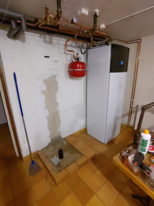 Rum med golv av klinker, en vit panna, rörinstallationer och en upphöjd plattform. En kvast står lutad mot väggen.
