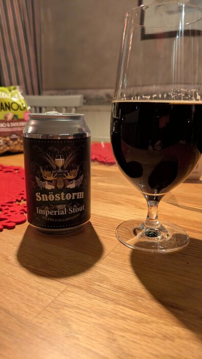 Burk och glas med Snöstorm Imperial Stout öl på träbord, omgiven av snacks.