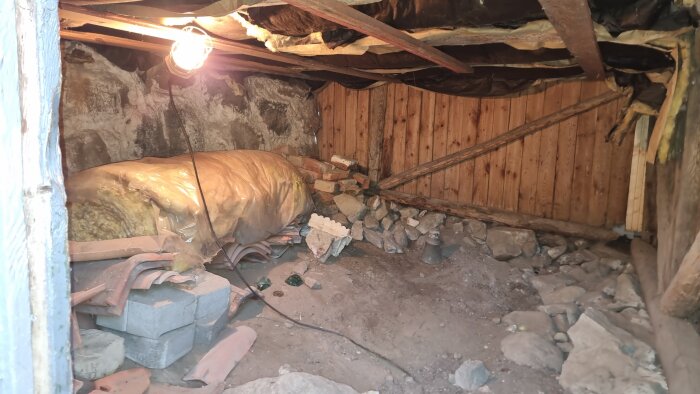 Krypgrund med stenblock, fuktig isolering och träväggar. Rötskador och staplade tegelstenar syns tydligt under varm lampa.