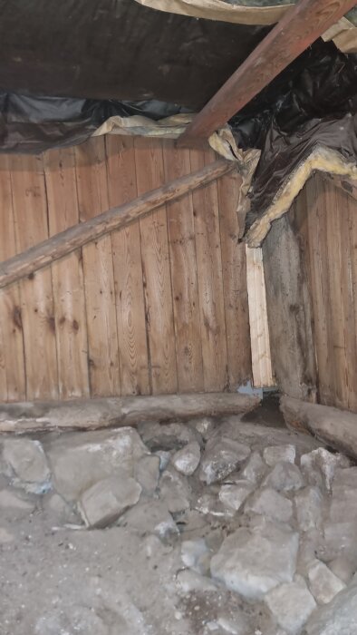 Rötskador och fuktfläckar i trästomme vid husgrund, omgiven av stenar och tegel i ett äldre hus från 1899.