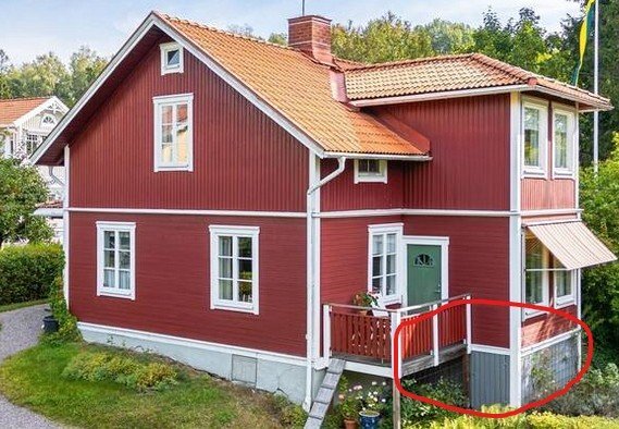 Rött trähus med vita knutar och tegeltak, omgiven av grönska. En del av altan markerad med röd cirkel. Trädgård med gräsmatta och buskar.