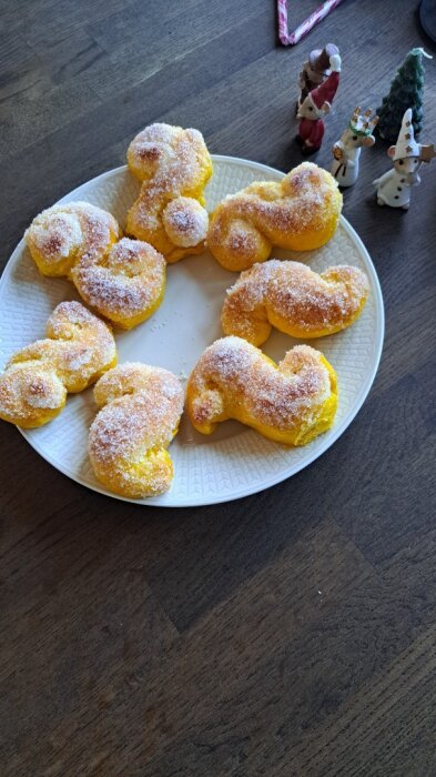 Gula lussekatter med socker på ett vitt fat, omgivna av små julfigurer på ett träbord.