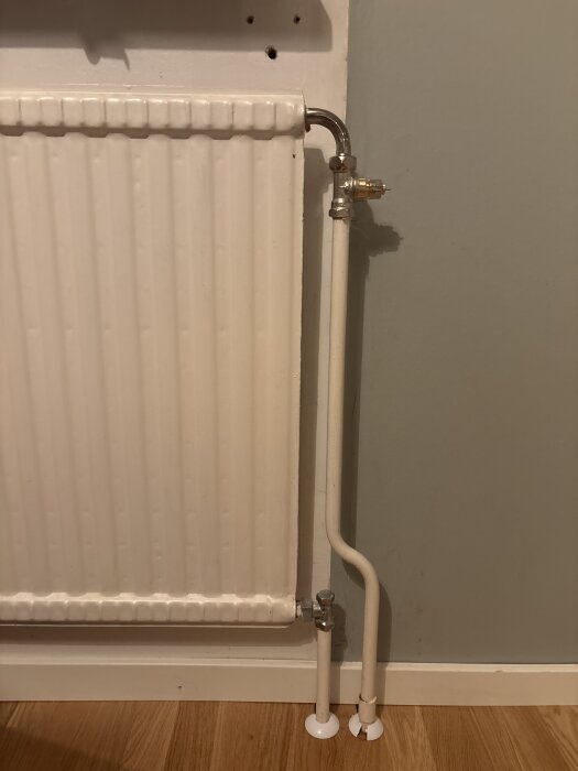 Vitt väggmonterat element med synliga rör och ventiler mot en grå vägg, stående på ett trägolv.