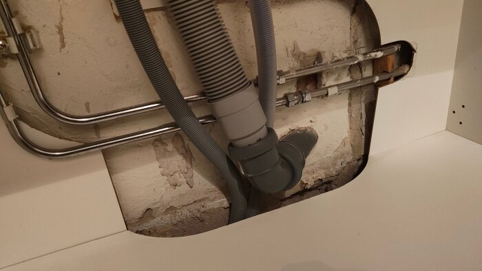 Rör och avlopp synliga under diskbänken i ett äldre kök, ett plaströr och metallrör mot en putstig vägg.