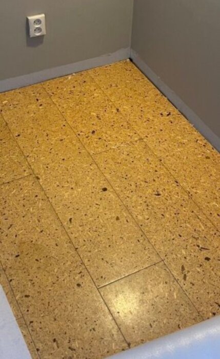 Golvet i hallen är täckt av beige-bruna plattor, eventuellt plast eller kork/linoleum, med eluttag synligt på väggen.