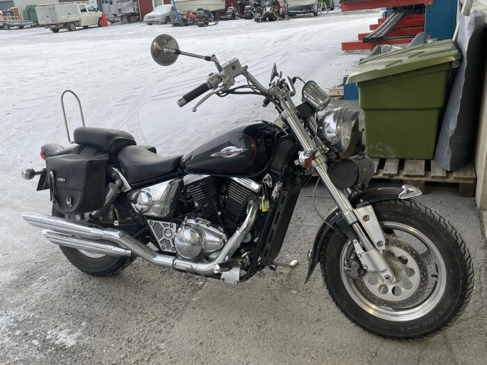 Svart Suzuki Marauder motorcykel parkerad på snötäckt mark, bredvid en grön soptunna och med hangar i bakgrunden.