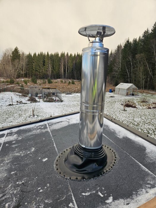 Silverfärgad skorsten monterad på ett tak med snö omkring, omgiven av skog och byggnader i bakgrunden.