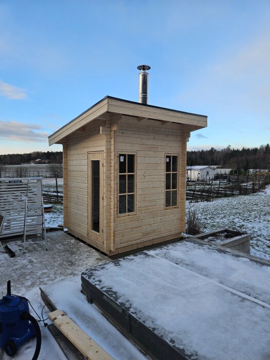 Liten, nybyggd bastustuga i trä med skorsten, omgiven av snö och landskap.