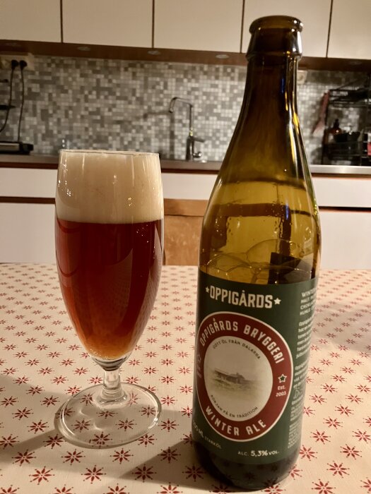 Flaska och glas med Oppigårds Winter Ale på bord med stjärnmönstrad duk, i kök med kakelvägg och diskbänk i bakgrunden.