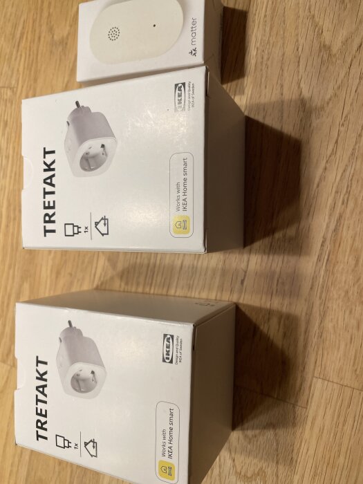 IKEA TreTakt smartpluggar och en Matter-enhet i oöppnade kartonger, liggande på ett trägolv.