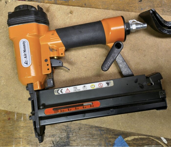 Orange och svart tryckluftsklammerpistol med etikett "Air Mounty" ligger på en träyta.