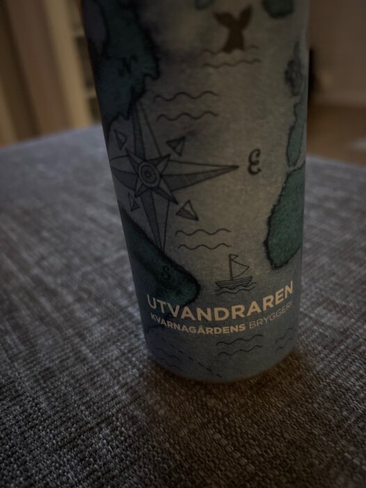 Flaska med karta och texten "UTVANDRAREN KVARNAGÅRDENS BRYGGERI" på etiketten, ståendes på en grå yta.