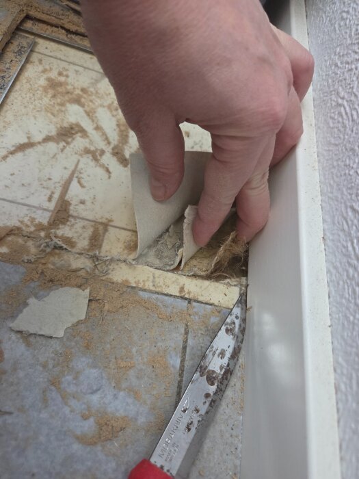Hand drar upp golv som visar flera lager, inklusive laminat och plastmatta, med en kniv vid sidan.