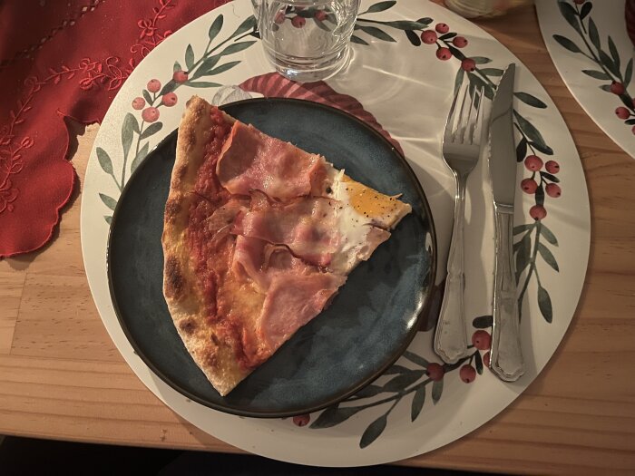En skiva pizza med skinka och ägg på en tallrik med blad- och bärmotiv, serverad med glas, gaffel och kniv.