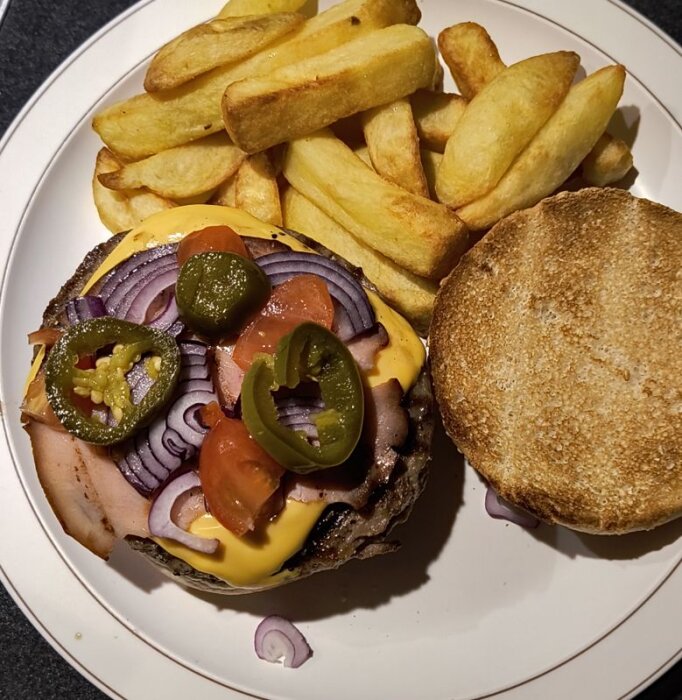 Hamburgare med ost, skinka, rödlök, tomat och jalapenos på tallrik med pommes.