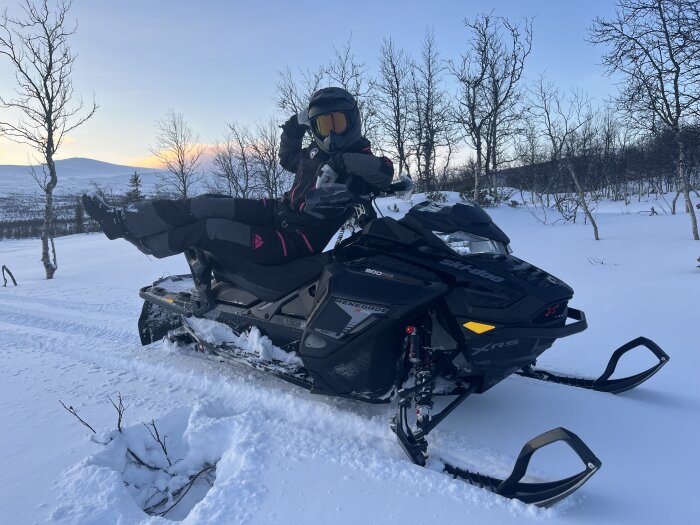 Person kör snöskoter i vintrigt landskap, iklädd hjälm och vinterkläder, med snöklädda träd och fjäll i bakgrunden under solnedgången.
