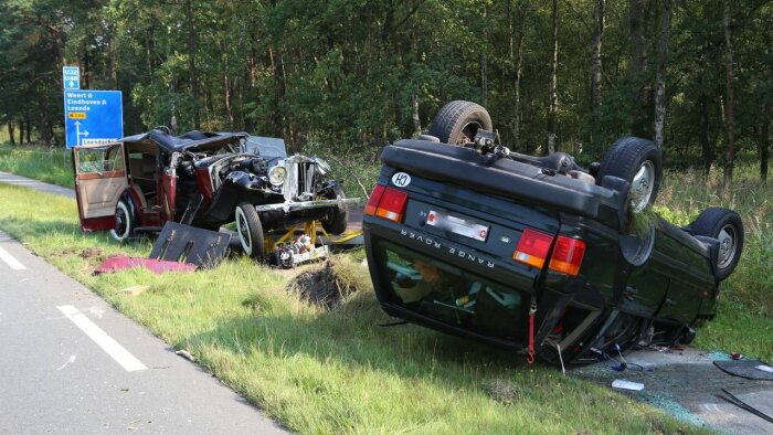 Krockad bil och vält Range Rover vid vägkant med skylt mot Weert och Eindhoven i bakgrunden, påminner om vikten av rätt lastning.