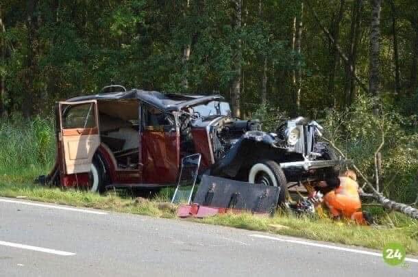 Kraschad gammaldags bil vid vägkant, trasig kaross och öppna dörrar. Skog i bakgrunden, person i orange overall undersöker vraket.