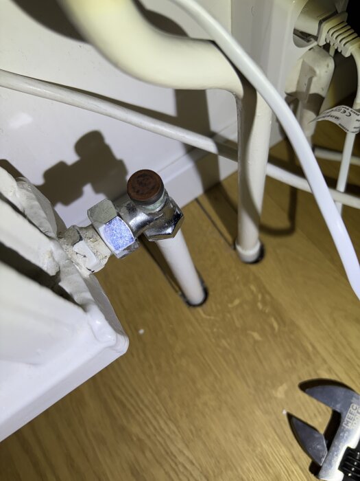 En rostig ventil med rör vid en vit radiator, inomhusmiljö på trägolv. En skiftnyckel syns också.