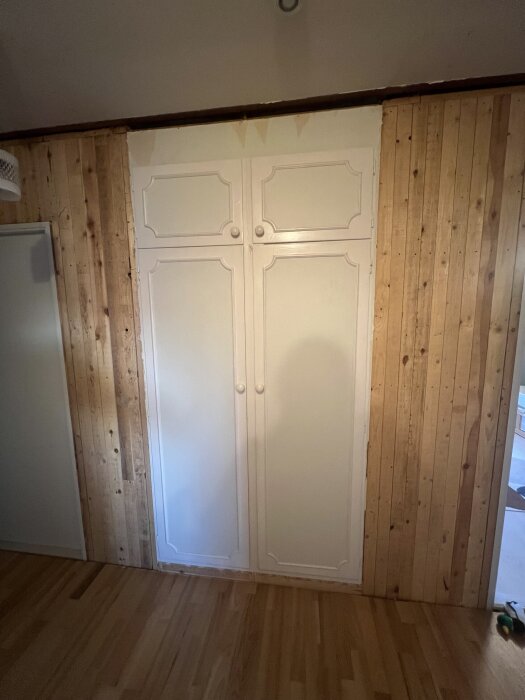 Inbyggda 60-tals garderober med vita dörrar i en hall under renovering, omgivna av brädvägg och trägolv.