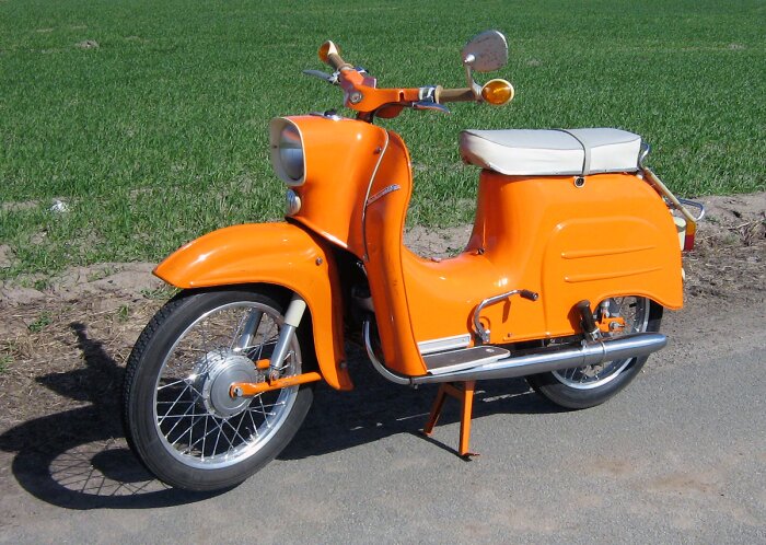 Orange moped parkerad på landsväg med grönt fält i bakgrunden.