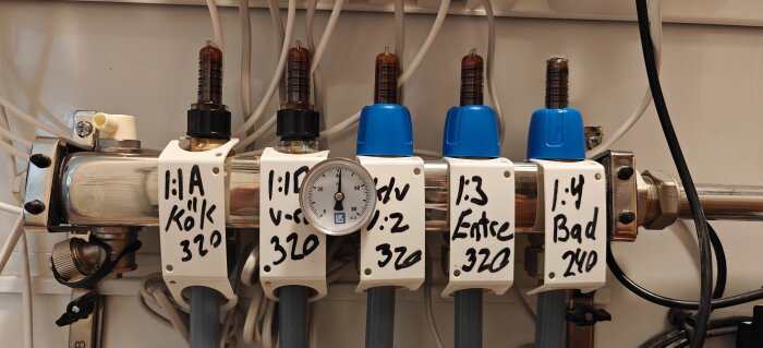 Manifold system with labeled flow meters and a pressure gauge for a heating system, showing various zones such as kitchen, entrance, and bathroom.